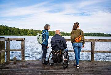 Ein Mann im Rollstuhl und zwei Frauen stehen auf einem Holzsteg am See und blicken auf das Wasser. Die Personen sind von hinten zu sehen, die Frauen stehen rechts und links von dem Mann und tragen jeweils einen Rucksack beziehungsweise Umhängetasche.