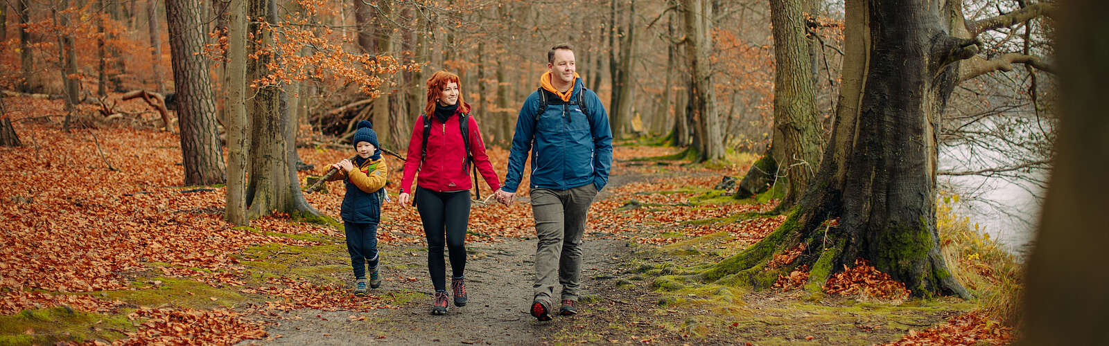 Eine Familie aus Vater, Mutter und Kind wandert durch einen herbstlichen Wald am Seeufer