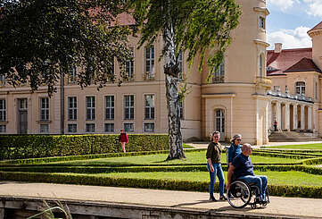 Ein Mann im Rollstuhl und zwei Frauen spazieren durch den angelegten Park vor dem Schloss Rheinsberg.