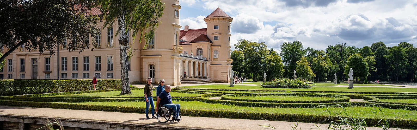 Ein Mann im Rollstuhl und zwei Frauen spazieren durch den angelegten Park vor dem Schloss Rheinsberg.