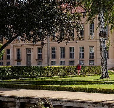 Ein Mann im Rollstuhl und zwei Frauen spazieren durch den angelegten Park vor dem Schloss Rheinsberg.