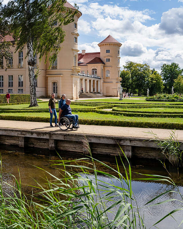Ein Mann im Rollstuhl und zwei Frauen spazieren durch den angelegten Park vor dem Schloss Rheinsberg.