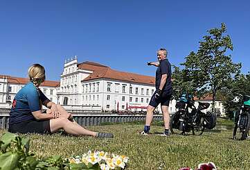 Eine Radfahrerin sitzt auf einer Wiese und hält eine Radkarte in der Hand, ein Radfahrer steht neben ihr und schaut auf das hinter ihnen liegende Schloss Oranienburg. Neben ihnen stehen die Fahrräder mit Gepäcktaschen.