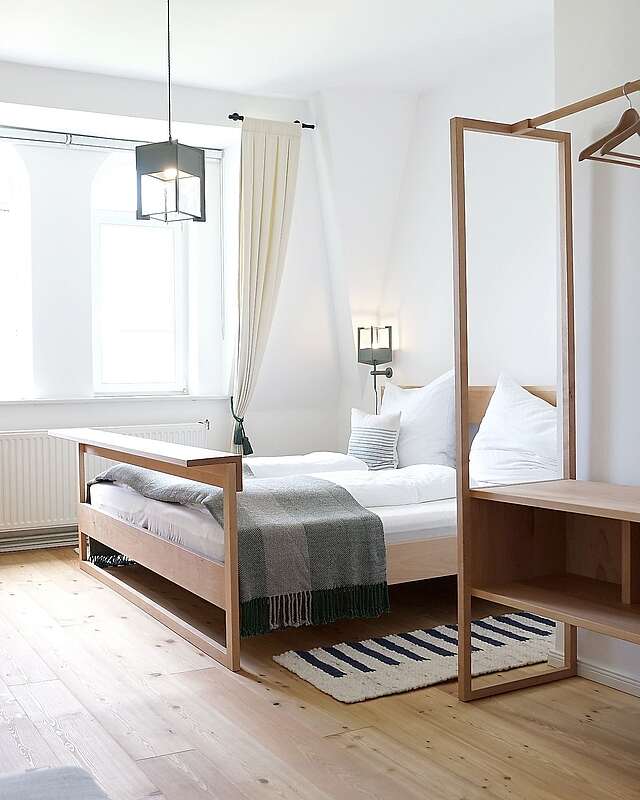Moderne Schlafzimmermöbel aus hellem Holz stehen in einem lichtdurchfluteten Zimmer. Cremefarbene Vorhänge und der Holzfußboden tragen zur Natürlichkeit bei.
