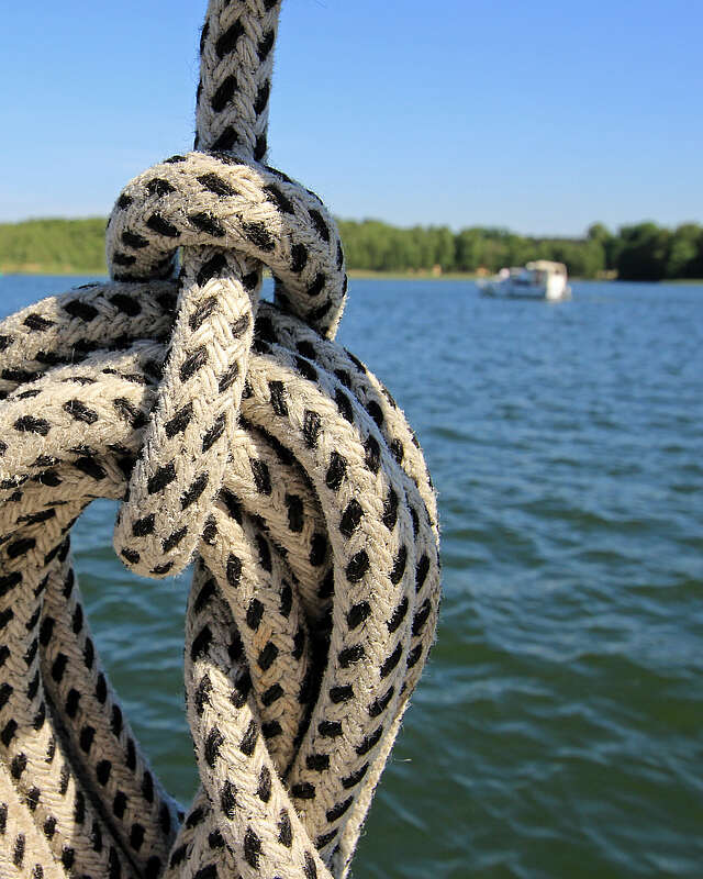 Ein Seemannsknoten in einem weißen Seil mit schwarzen Punkten. Im Hintergrund sieht man einen weiten See mit waldigem Ufer und mehrere Boote.