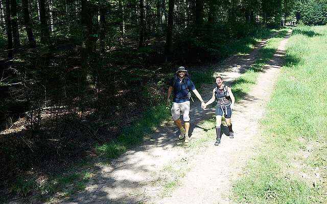 Ein Mann und eine Frau wandern in sommerlicher Outdoor-Kleidung Hand in Hand durch den Wald. Die Sonne scheint und beide lächeln in die Kamera.