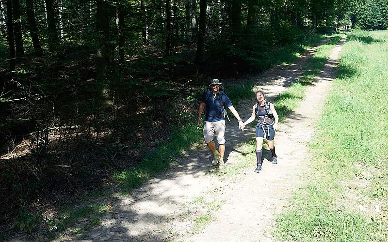 Ein Mann und eine Frau wandern in sommerlicher Outdoor-Kleidung Hand in Hand durch den Wald. Die Sonne scheint und beide lächeln in die Kamera.