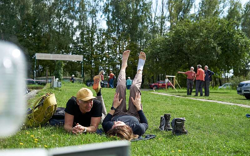 Ein Mann und eine Frau liegen in sportlicher Kleidung auf einer Wiese, die Beine in die Luft, und ruhen sich aus.