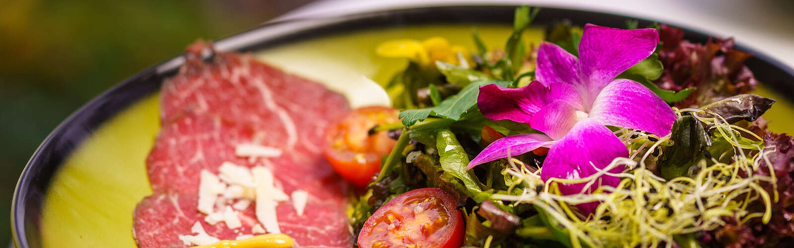 Fotos eines grünen, hübsch angerichteten Tellers mit roter Salami, kleinen Tomatenhälften, Salat und violetten Blüten.