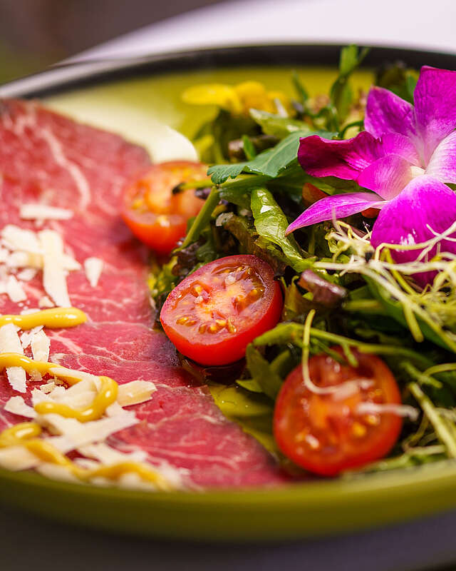 Fotos eines grünen, hübsch angerichteten Tellers mit roter Salami, kleinen Tomatenhälften, Salat und violetten Blüten.