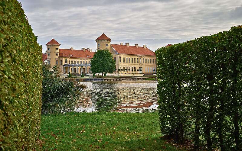 Schloss mit zwei runden Türmen hinter einem See, der Blick ist eingerahmt von hohen Hecken