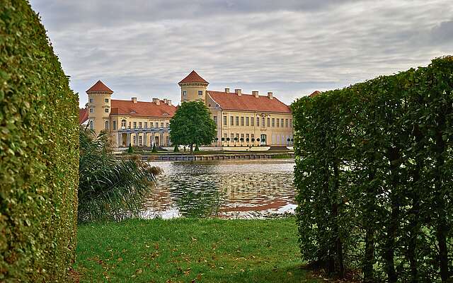 Schloss mit zwei runden Türmen hinter einem See, der Blick ist eingerahmt von hohen Hecken