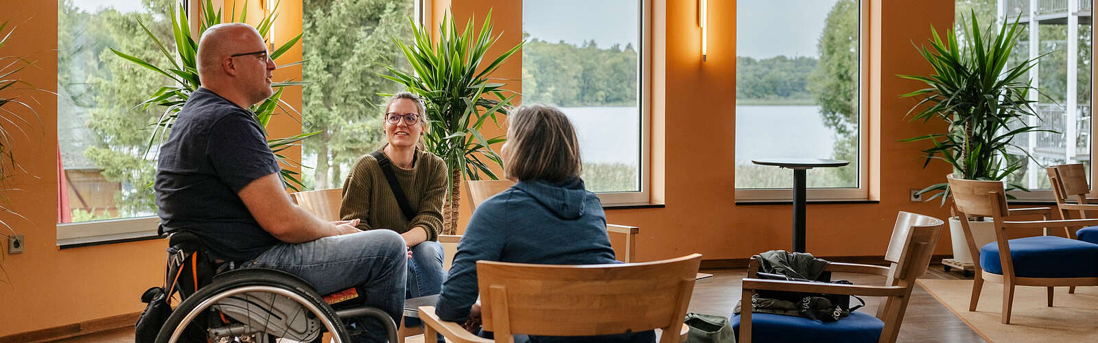 Ein Mann im Rollstuhl und zwei Frauen sitzen an einer Sitzgruppe im Foyer des Seehotel in Rheinsberg. Hinter ihnen kann man durch die großen Fenster den Grienericksee erkennen.
