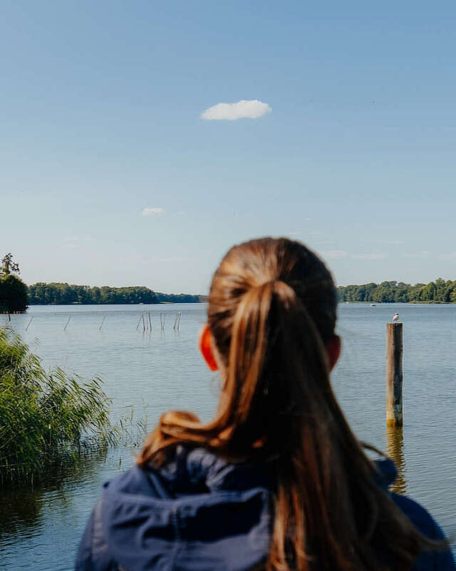Eine junge Frau steht an einem Geländer und schaut über den blauen See. Sie ist von hinten zu sehen.