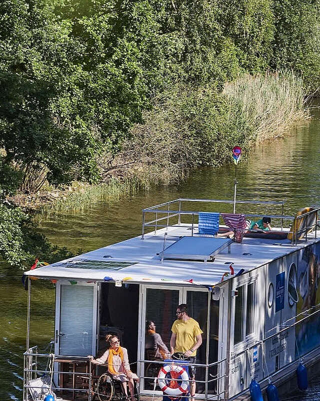 Ein Hausboot fährt in einen Kanal. Ein Mann steht hinter dem Steuer, eine Frau sitzt neben ihm im Rollstuhl. Oben auf dem Sonnendeck liegt ein Junge und liest.