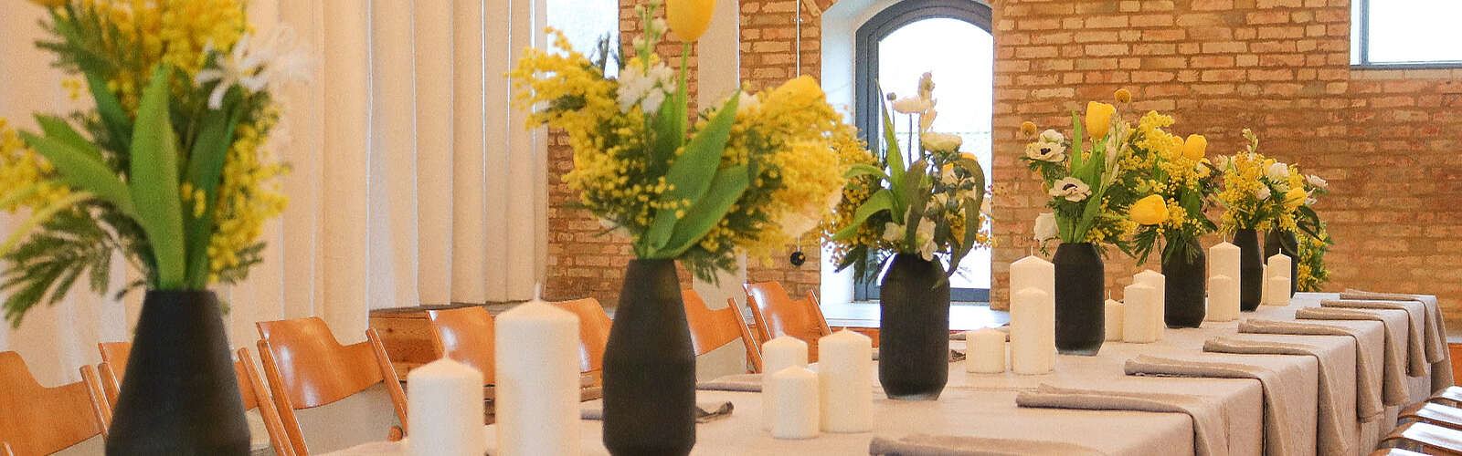 Ein hübsch dekorierter Tisch vor einer Steinwand. Auf der hellen Tischdecke stehen schwarze Vasen mit gelben Blumen. Kerzen und Stoffservietten runden das Gesamtbild ab.