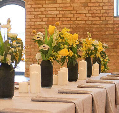Ein hübsch dekorierter Tisch vor einer Steinwand. Auf der hellen Tischdecke stehen schwarze Vasen mit gelben Blumen. Kerzen und Stoffservietten runden das Gesamtbild ab.