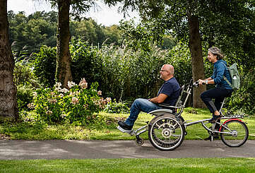 Ein Mann und eine Frau fahren gemeinsam auf dem Rollfiets über einen asphaltierten Weg, der auf der einen Seite von Uferschilf gesäumt wird. Die Frau tritt in die Pedale, der Mann hat auf dem vorderen Sitz Platz genommen.