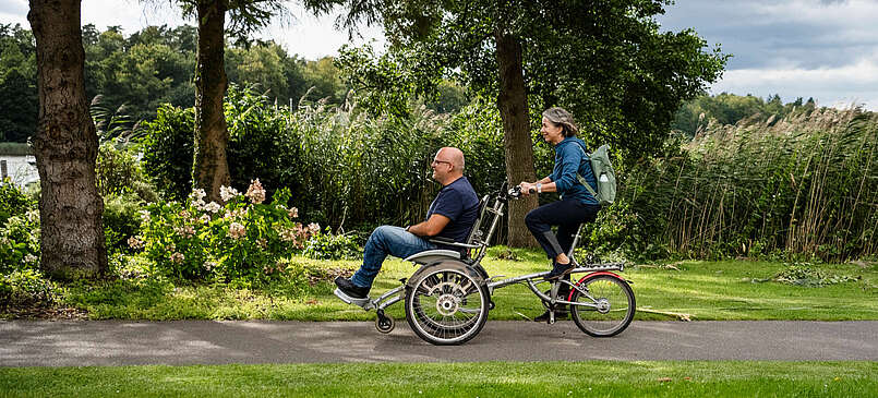 Ein Mann und eine Frau fahren gemeinsam auf dem Rollfiets über einen asphaltierten Weg, der auf der einen Seite von Uferschilf gesäumt wird. Die Frau tritt in die Pedale, der Mann hat auf dem vorderen Sitz Platz genommen.