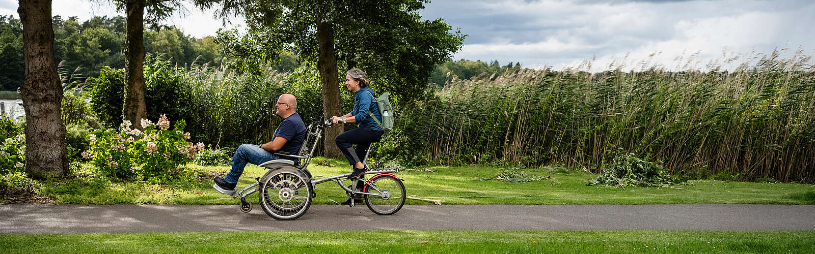 Ein Mann und eine Frau fahren gemeinsam auf dem Rollfiets über einen asphaltierten Weg, der auf der einen Seite von Uferschilf gesäumt wird. Die Frau tritt in die Pedale, der Mann hat auf dem vorderen Sitz Platz genommen.