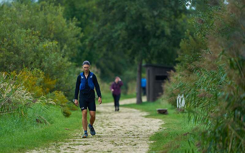 Ein sportlich gekleideter junger Mann wandert auf einem leicht sandigen Weg entlang von Schilf und Bäumen.