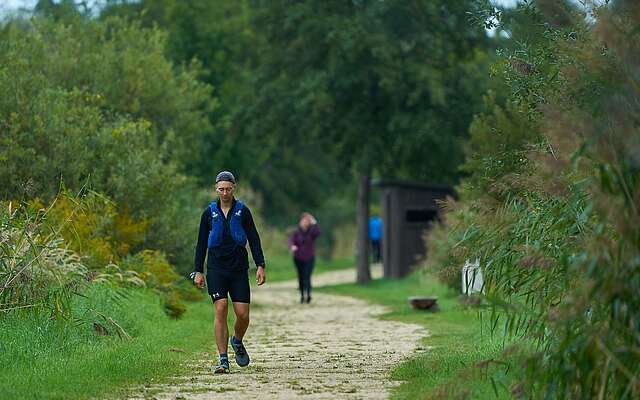 Ein sportlich gekleideter junger Mann wandert auf einem leicht sandigen Weg entlang von Schilf und Bäumen.