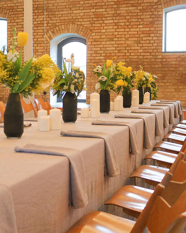 Ein hübsch dekorierter Tisch vor einer Steinwand. Auf der hellen Tischdecke stehen schwarze Vasen mit gelben Blumen. Kerzen und Stoffservietten runden das Gesamtbild ab.