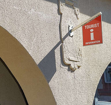 Eine Häuserfront mit oben abgerundeten Fenstern. An der Scheibe und an der verputzten Wand hängen Schilder mit dem roten "i" und eine Lampe.