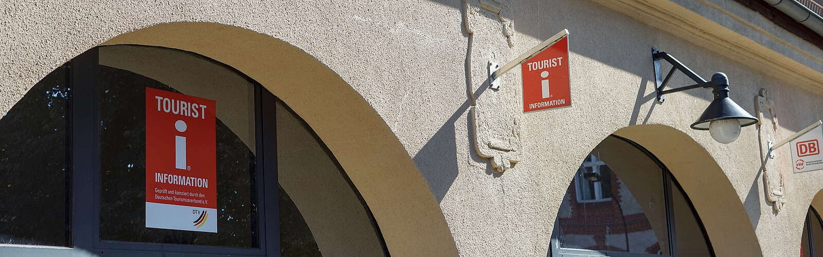 Eine Häuserfront mit oben abgerundeten Fenstern. An der Scheibe und an der verputzten Wand hängen Schilder mit dem roten "i" und eine Lampe.
