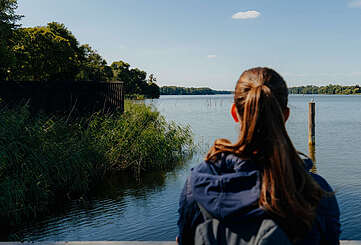 Eine junge Frau steht an einem Geländer und schaut über den blauen See. Sie ist von hinten zu sehen.