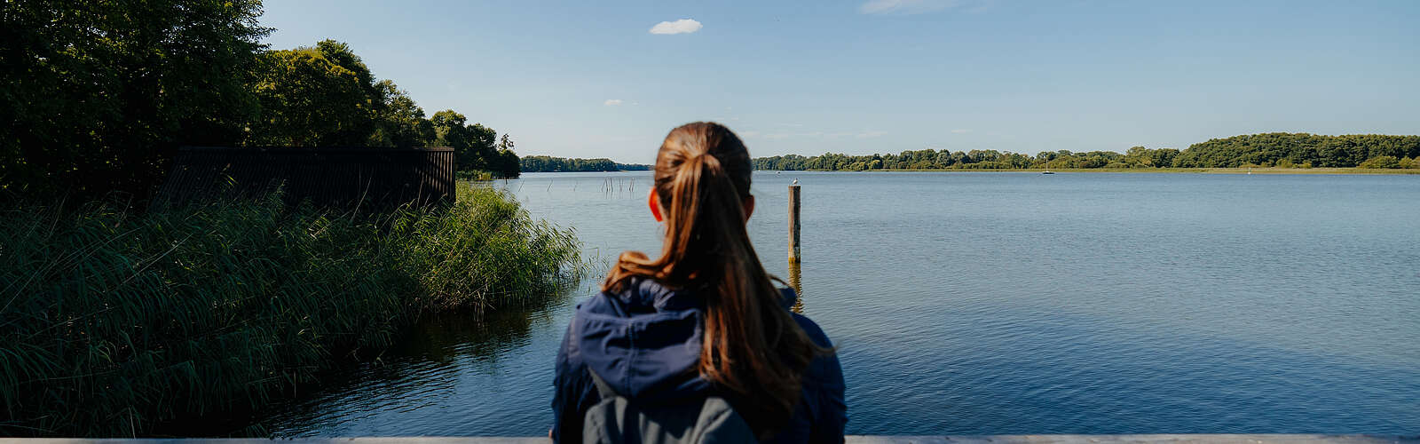 Eine junge Frau steht an einem Geländer und schaut über den blauen See. Sie ist von hinten zu sehen.