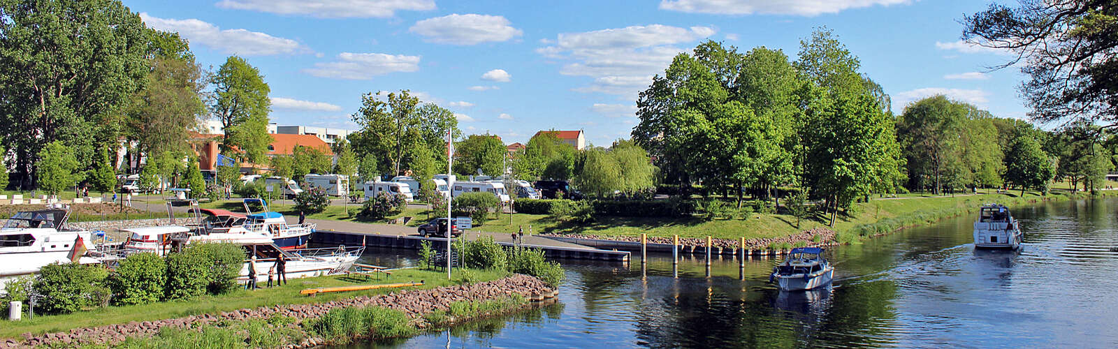 Boote fahren bei sommerlichem Wetter auf einem Kanal. Nach links führt eine kleine Wasserstraße zu Anlegeplätzen. Im Hintergrund sind Wohnmobile an Land zu erkennen.