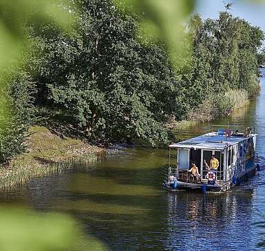 Ein Hausboot fährt in einen Kanal. Ein Mann steht hinter dem Steuer, eine Frau sitzt neben ihm im Rollstuhl. Oben auf dem Sonnendeck liegt ein Junge und liest.