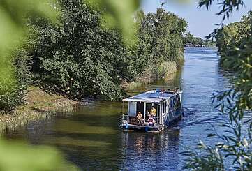 Ein Hausboot fährt in einen Kanal. Ein Mann steht hinter dem Steuer, eine Frau sitzt neben ihm im Rollstuhl. Oben auf dem Sonnendeck liegt ein Junge und liest.